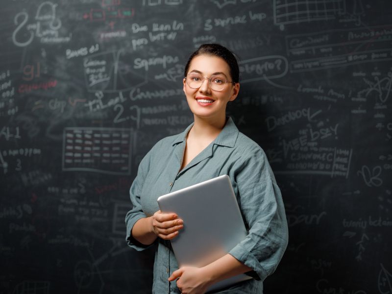 Profesora con portátil en la mano delante de la pizarra de clase.